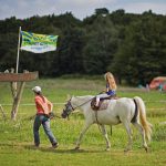 Een wit paard met een meisje erop en een vrouw ernaast, wandelend over de camping