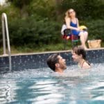 Een man met kind aan het spelen in het zwembad van de natuurcamping met een vrouw die toekijkt