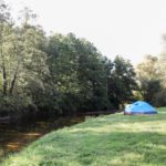 Een blauwe tent staat op het gras, direct aan de over van de rivier.