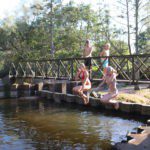 Twee kinderen springen met opgetrokken knieën van een bruggetje in de rivier.