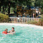 Zwembad met daarachter een speeltuin op Camping Huttopia Lac de Sillé